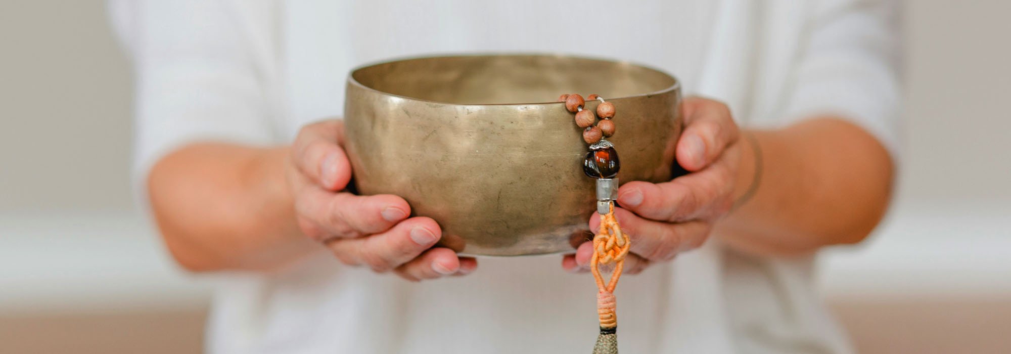 Two hands cradling an aged Tibetan singing bowl with a wooden mala draped over the rim, against a soft white background