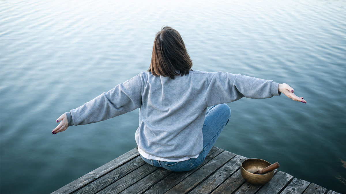 Meditating outdoors by water with a Tibetan singing bowl Woman meditating cross-legged on a lakeside dock with a Tibetan singing bowl and mallet beside her