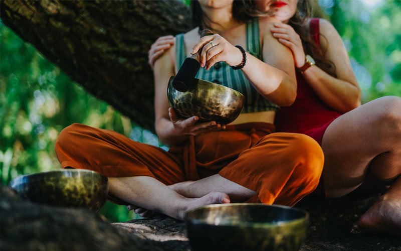 Two close female friends playing a handmade Tibetan singing bowl together in nature for meditation, friendship, and sound healing
