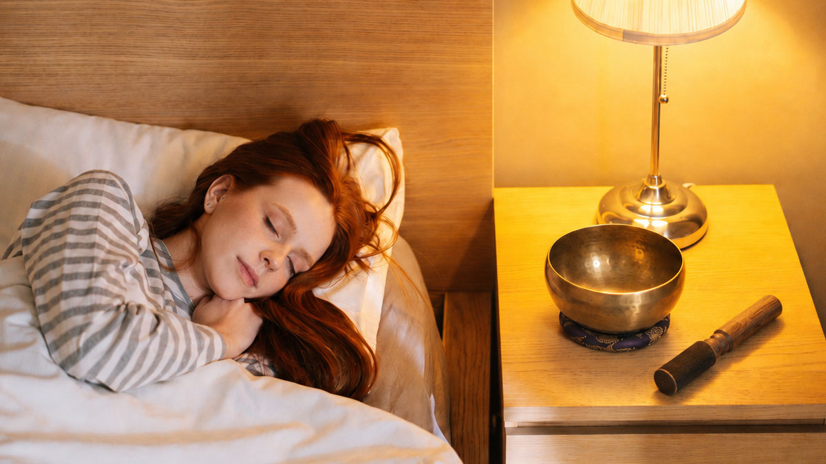 Singing bowl for sleep — kept bedside for an evening wind-down practice Tibetan singing bowl and mallet on a bedside table beside a woman sleeping peacefully