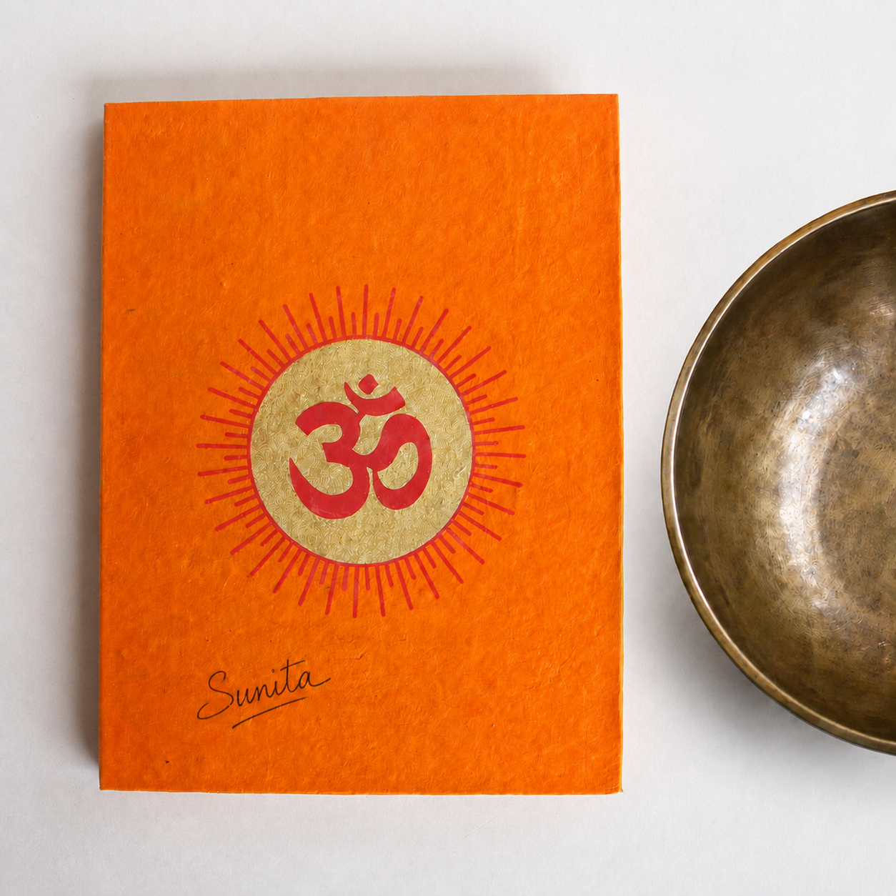 Sunita Poudel's handwritten sound healing training notebook, orange lokta paper with a gold and red Om, beside a hand-hammered singing bowl
